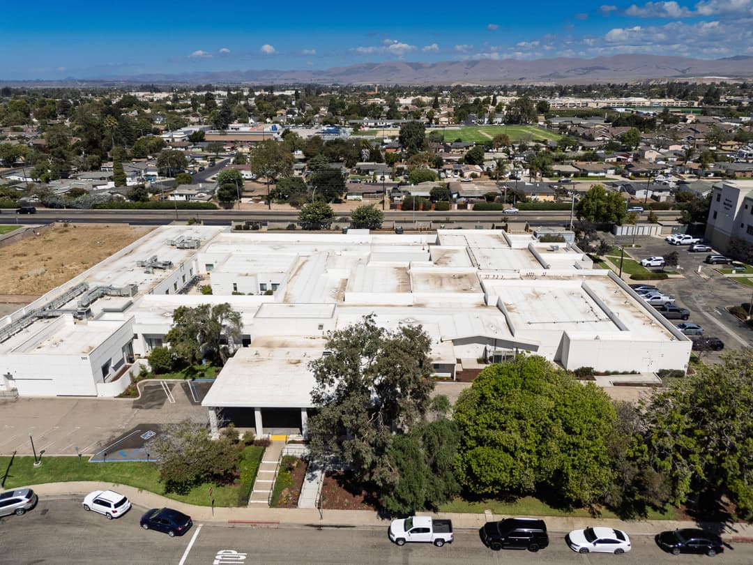 Aerial view of a modern building surrounded by parking and green spaces in an urban landscape.