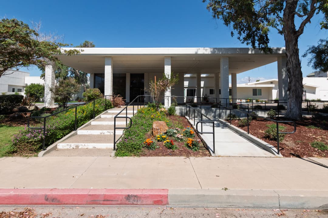 Modern building entrance with landscaped garden and accessible ramp, clear blue sky above.