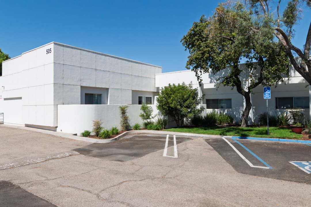 Modern white building with ample landscaping and parking spaces under a clear blue sky.