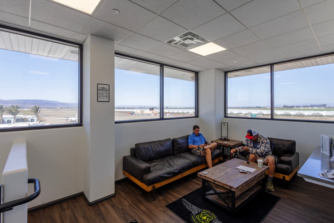 Furnished waiting area with two men, large windows showing airport runway and landscape.