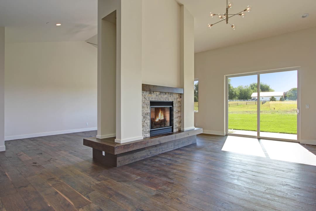 Modern living room with stone fireplace, hardwood floors, and large windows overlooking a green landscape.