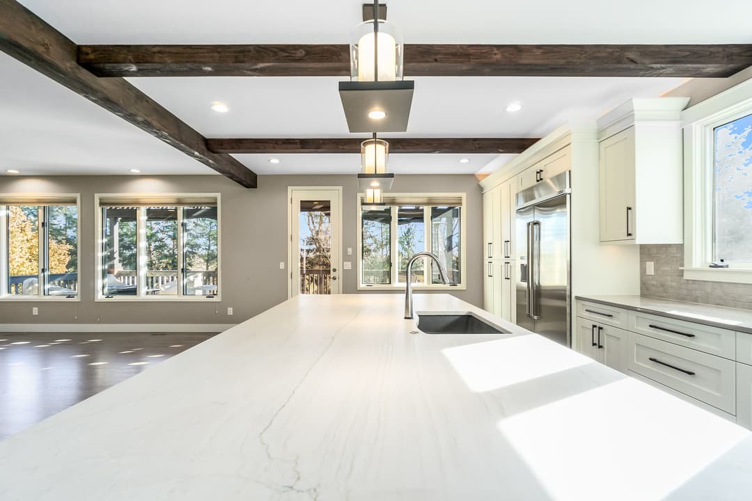 Modern kitchen with white marble island, wooden beams, and large windows for natural light.