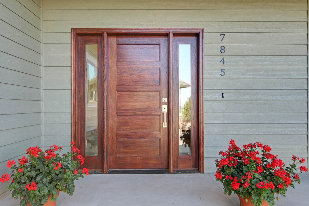 Modern wooden front door with sidelights, surrounded by red flowers, at 7845 address.