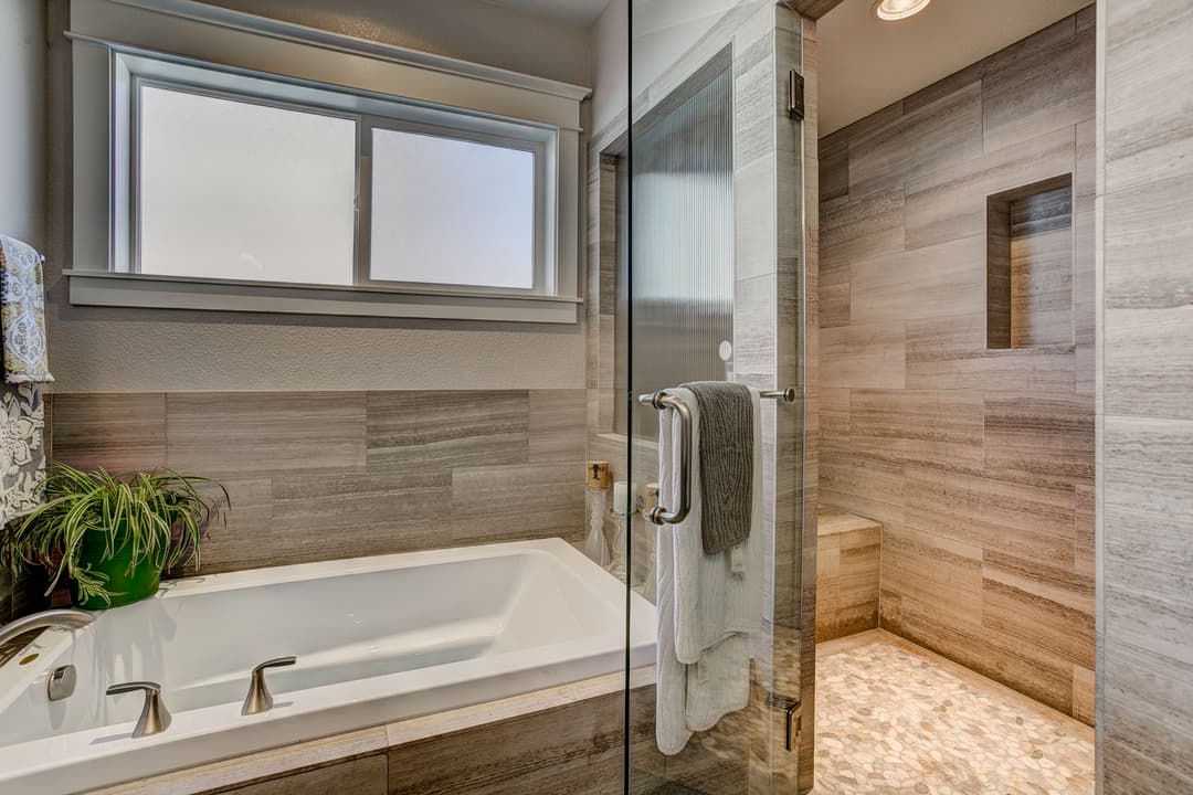 Modern bathroom with a glass shower, white bathtub, and gray tile accents, featuring natural light.