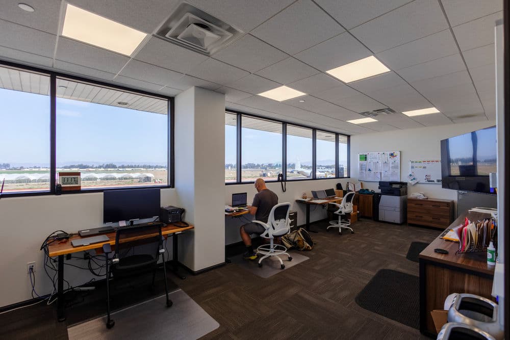 Modern office workspace with desks, computers, and large windows overlooking a landscape.